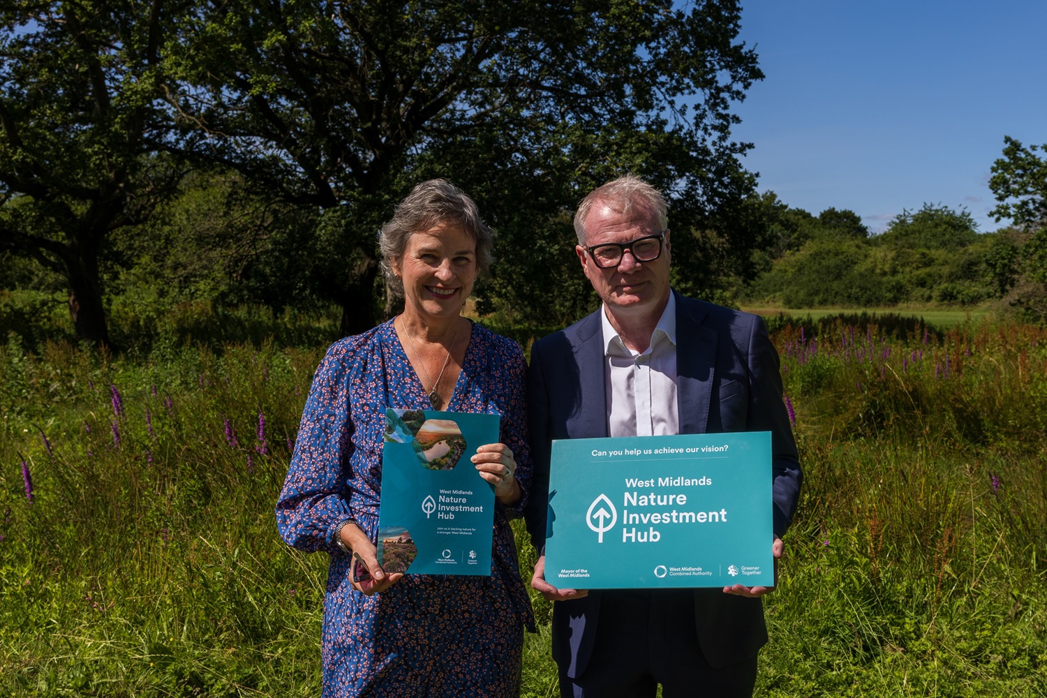 Two people stand in a sunny field holding "West Midlands Nature Investment Hub" signs, smiling, with greenery and trees in the background.