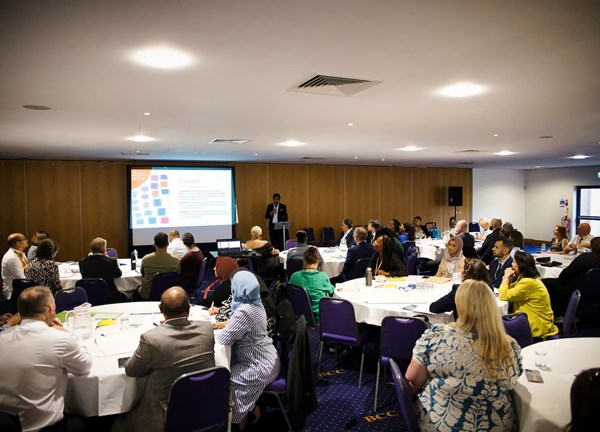 Conference room with multiple round tables where attendees are seated, listening to a speaker presenting slides on a large screen at the front