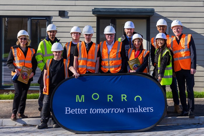 Group of 11 people in bright-orange hi-vis vests and white hard-hats standing behind a blue banner bearing the name Morro.