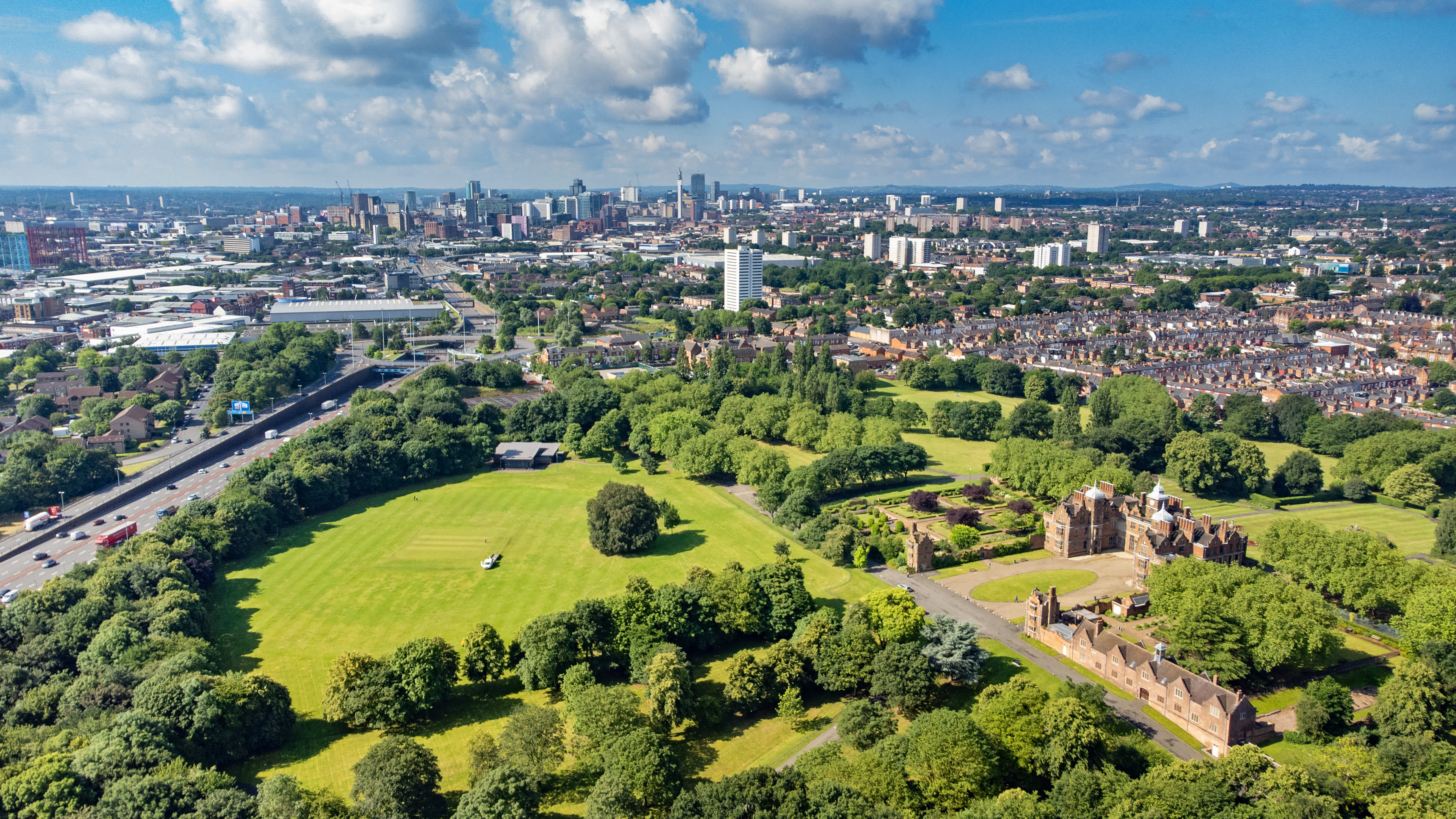 Aerial view of Birmingham with a large green park, historic red-brick building, and city skyline under a blue sky