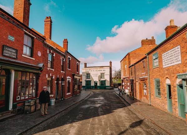 A landscape photograph featuring a High Street located at Dudley's Black Country Living Museum. In the scene, two actors are standing outside their respective shops.