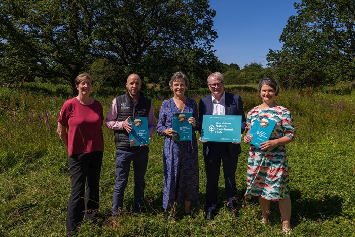 Five people stand outdoors in a grassy field, holding signs and booklets with trees and blue sky in the background.