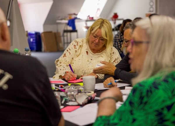 People sit around a table in a bright indoor workspace, collaborating on a hands‑on activity. Sticky notes, pens, paper, a coffee cup, and glasses are scattered across the table. One person writes on a pink sticky note while another gestures beside them, suggesting active discussion and teamwork.