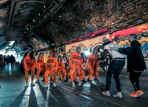A photograph of a troupe of dancers showcasing a choreographed piece which is being recorded by a videographer. They are dancing in a fashionable, graffitied tunnel which is a public space.