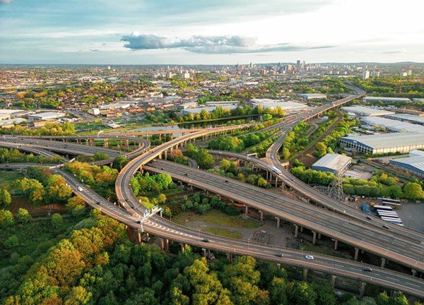 Aerial view of a multi-level motorway interchange surrounded by green trees, industrial buildings, and a distant city skyline under a partly cloudy sky.