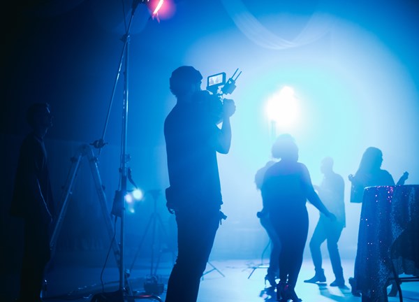 A photograph capturing a behind-the-scenes film shoot. The blue lighting in the room cuts through the dark, foggy atmosphere of unknown actors dancing in the scene.