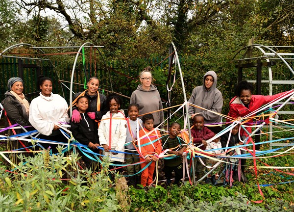 A group portrait of the Glue Collective posing with families and children outdoors in a green area reminiscent of an allotment.