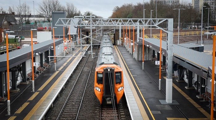 A train with yellow front in a new station with platforms either side, canopies and in the distance a footbride