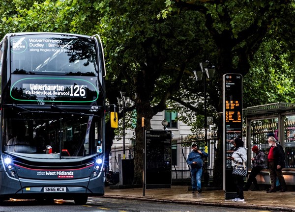 A dark grey National Express West Midlands "Platinum" double-decker bus, displaying route "126 to Wolverhampton via Hagley Road," approaches a bus stop. Passengers stand waiting under large green trees beside an electronic timetable display and a glass bus shelter.