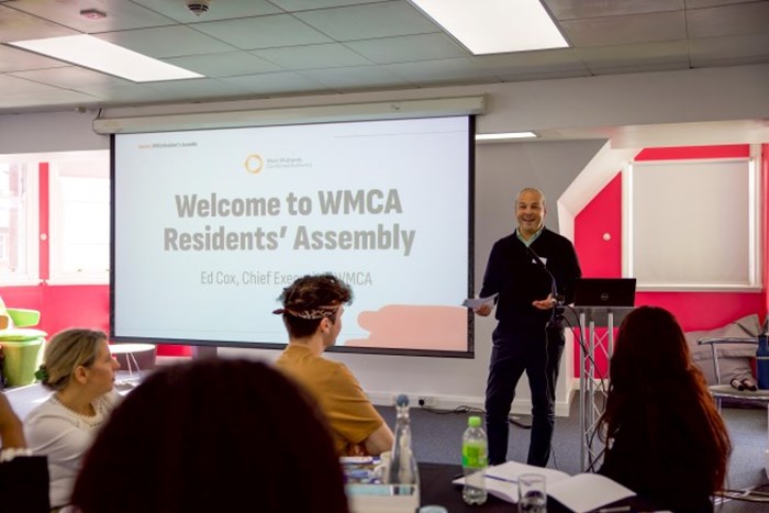 A man stands at a podium in a bright, modern conference room, speaking to seated attendees. Behind him, a large projection screen displays a slide that reads “Welcome to WMCA Residents’ Assembly – Ed Cox, Chief Executive, WMCA.” The room features red and white walls, natural light from tall windows, and tables with bottles and papers.