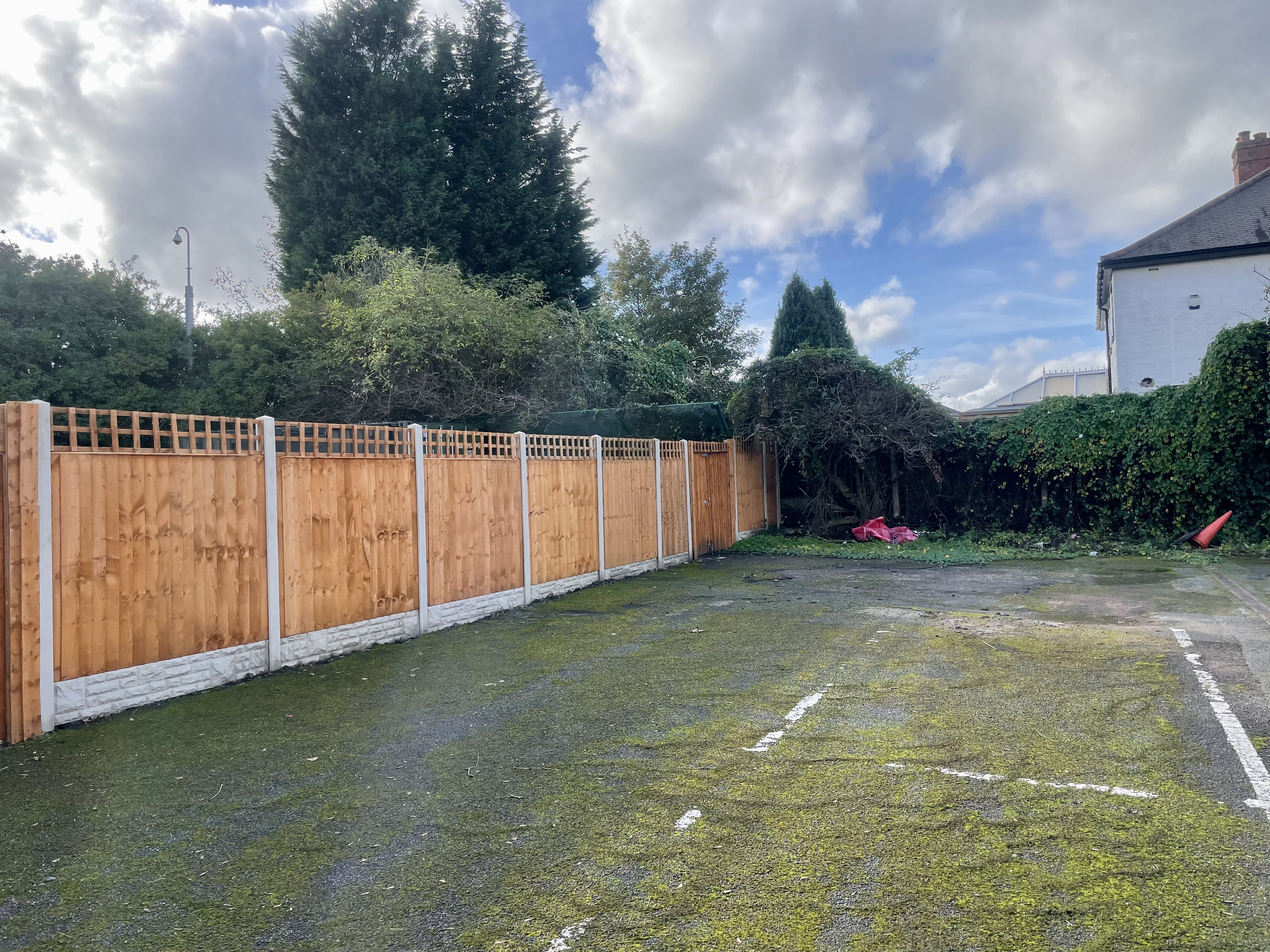 Image shows a disused car park with a fence around the side and unkept bushes at the end