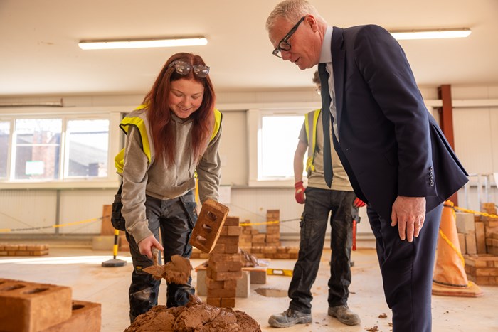 A woman shows a man how to lay a brick.