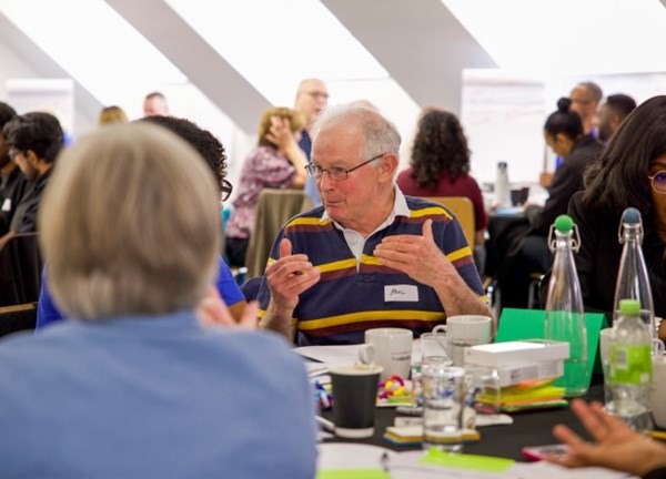 An older resident speaks and gestures with their hands while taking part in a small group discussion at the Residents' Assembly 2026. Participants of different ages and backgrounds are seated around tables with notebooks, drinks and workshop materials, while other groups can be seen talking in the background in a bright meeting space.