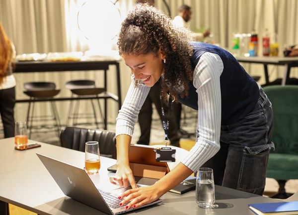 A young woman smiles as she leans over a desk, typing on a laptop. Glasses and books are scattered across the desk. In the background, people are chatting and socialising, with food and drinks on nearby tables, suggesting a networking or community event.