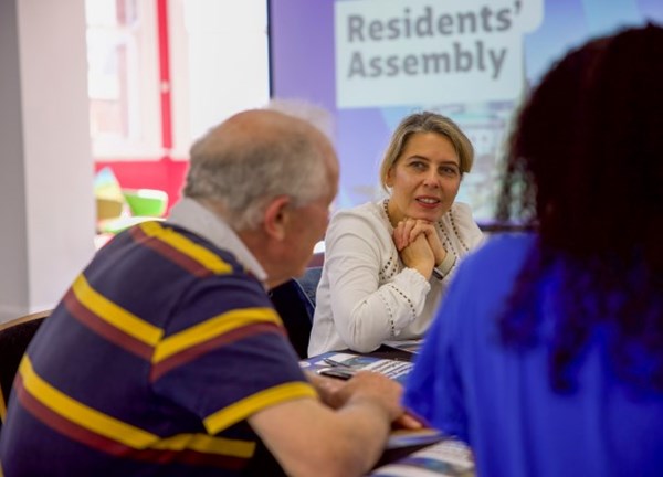 Three people sit around a table engaged in conversation during a community meeting. Behind them, a projected screen displays the words “Residents’ Assembly.” The setting appears informal and collaborative, with participants focused on discussion.