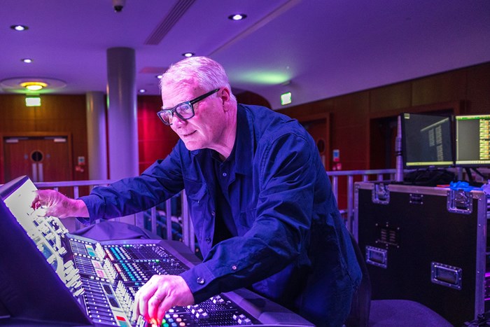A man with glasses and a blue shirt operates a professional audio mixing console with purple ambient lighting and monitors.