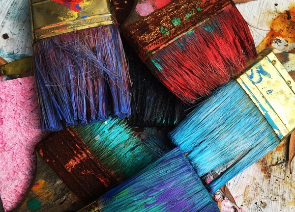 A colourful photograph featuring multiple used paintbrushes placed down on a workbench,