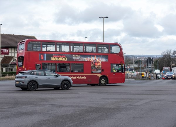 A red National Express West Midlands double-decker bus. The bus is driving alongside a grey crossover SUV across a wide paved junction, with a Toby Carvery building visible in the background under cloudy skies.