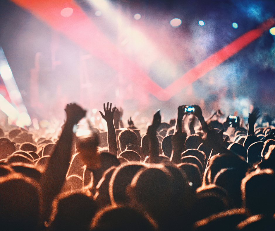 A large crowd cheering, looking towards the stage at a concert where there are bright lights.
