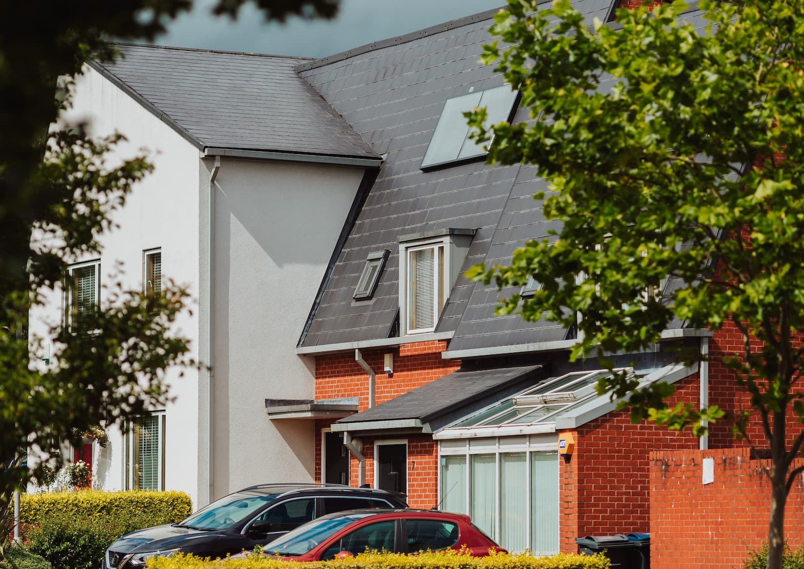 Modern semi-detached houses with sloped roofs, red brick and white walls, two parked cars in front, and green trees partially framing the view