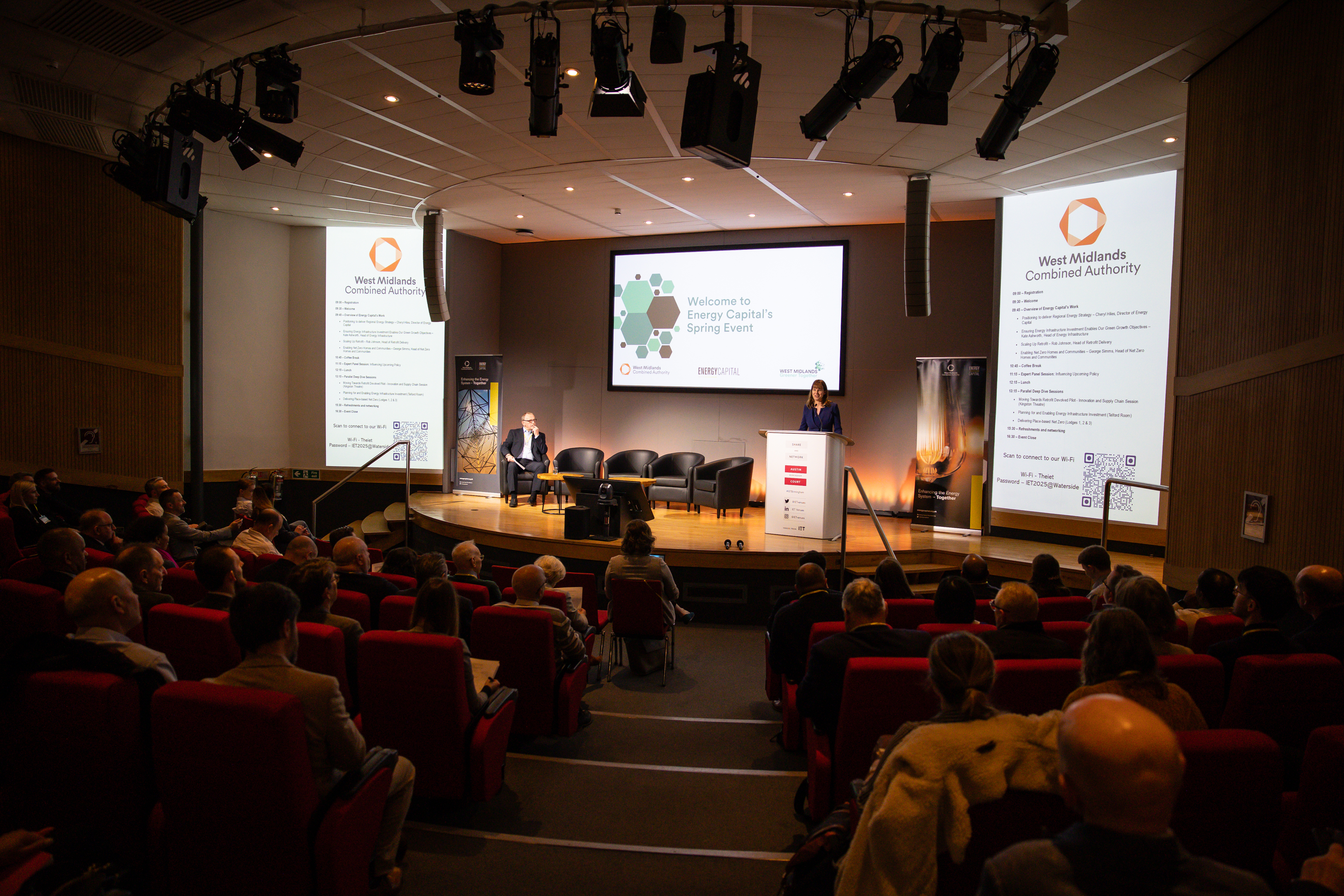 A lecture theatre full of people with someone presenting at a lectern on stage.
