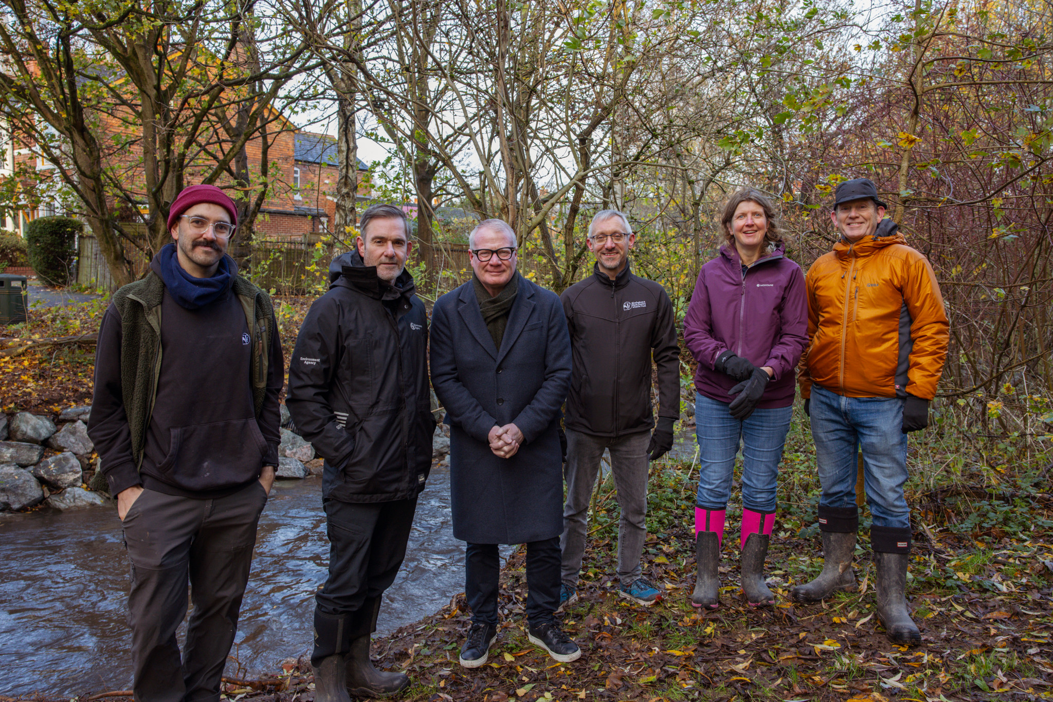 Six people in outdoor gear stand by a small stream in a wooded area. They appear content, with autumn leaves and a red brick building in the background.