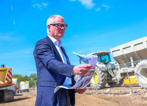 A man is pictured from the waist up standing in a suit. He is shown standing on a construction site. He is holding a construction site plan in his hands. He looks off to the right off camera. In the background are two heavy construction machines, include a track pulling a larger container to the right 