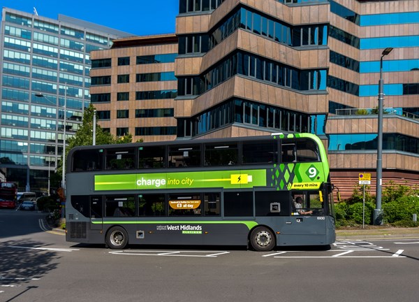 A grey and bright green electric double-decker bus operated by National Express West Midlands travelling through a city intersection. The bus features green branding along its side with the slogan "charge into city" and a battery icon, set against a backdrop of multi-story glass and brown office buildings under a clear blue sky.